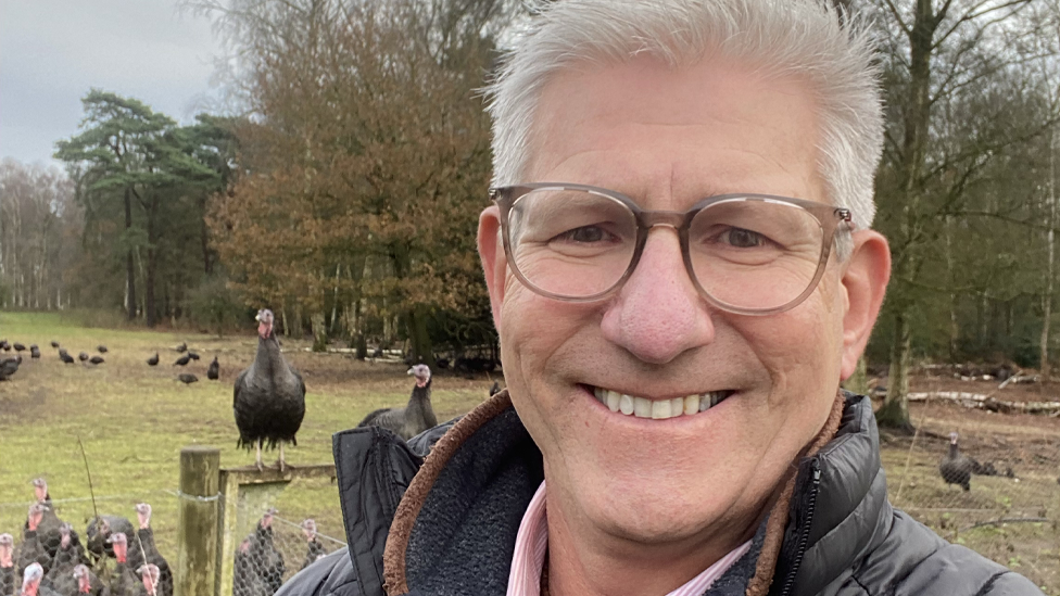 Mark Gorton smiling in a selfie with turkeys in a field behind him.