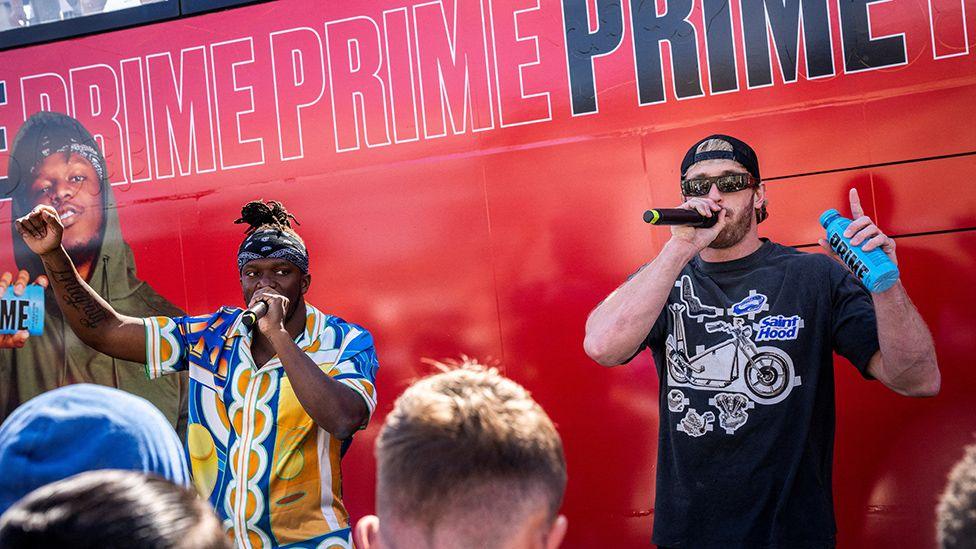 KSI and Logan Paul stand in front of a red bus marked repeatedly with the word "prime", apparently in front of a crowd (only a few heads visible). Both are talking through mics and Logan Paul is holding a blue bottle of Prime. KSI is wearing a blue and yellow Hawaiian shirt and a bandana; Paul is wearing a baseball cap back to front, and a black t-shirt with the insignia "Saint Hood"