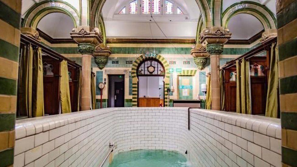 A highly decorated room with green and mustard tiles and ornate columns. There is a deep plunge pool in the foreground with white tiles. At the sides there are booths separated by wooden panels with decorations at the top and green privacy curtains.