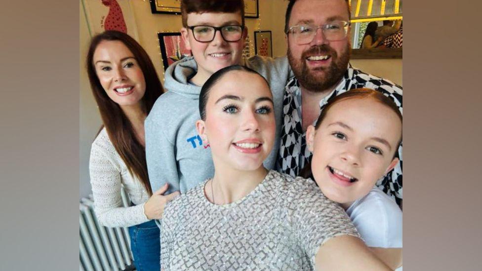 Joanne Kinsey is pictured with her husband and three children who are all smiling at the camera in a group selfie.