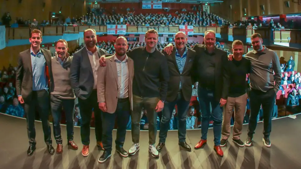 The Southend consortium: a group of men standing together shoulder to shoulder on a stage with a theatre of people behind them, and the England flag hanging from the balcony.