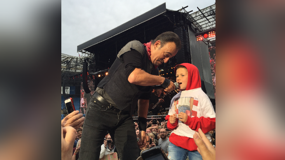 A young boy with a red hoody on, with the hood pulled on his head and a white t-shirt with is on stage singing with Bruce Springsteen. Springsteen is holding the microphone at the young boy, he is wearing black jeans and a black top. You can see the stage bhind and loads of fans in the photo behind and below the two who are on a stage.