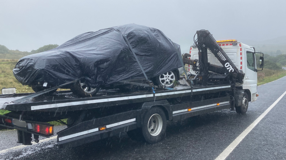 Car covered in black sheeting, on top of a removal lorry