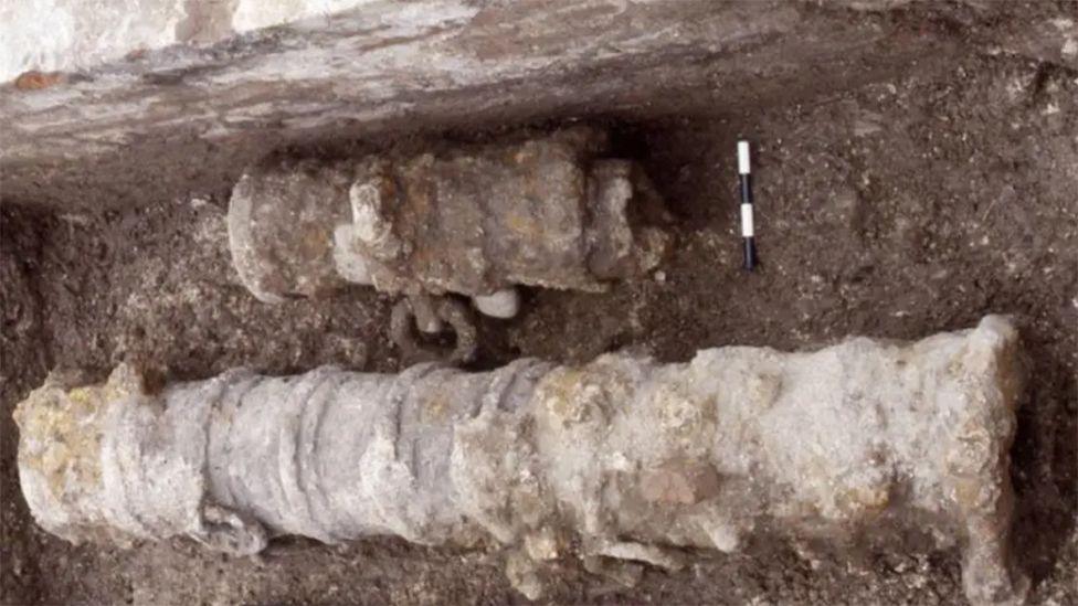 An archaeological photograph of a excavated cannon lying in a ditch which is in two parts and is covered in rust and earth