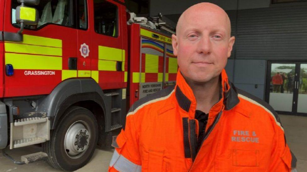 Jonathan Dyson stood in front of a fire engine wearing an orange and black jacket that says "fire and rescue" on it. He has a bald head and is looking at the camera with a slight smile.