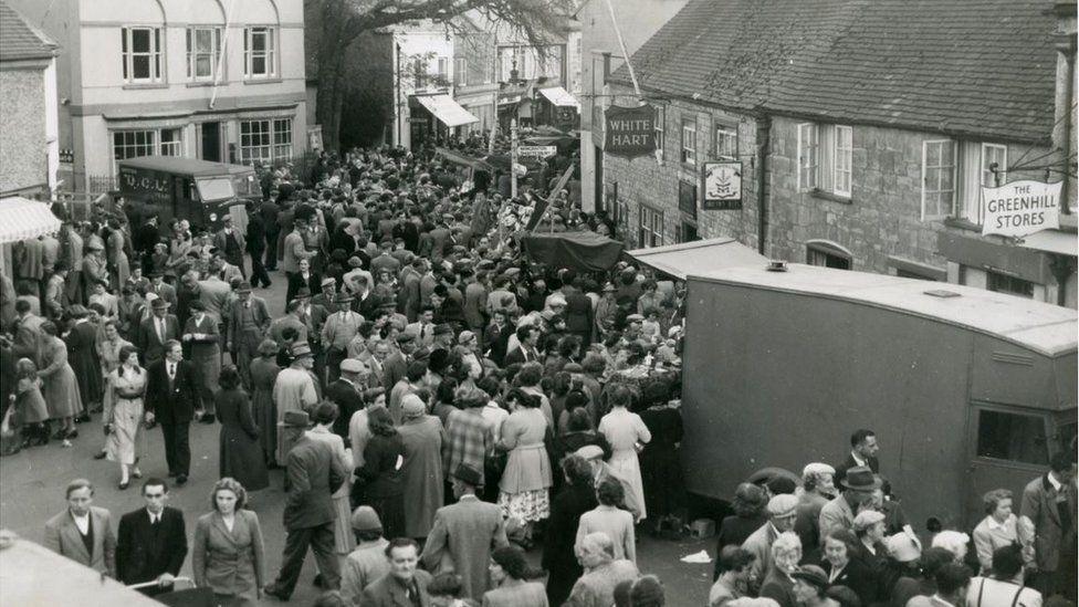 A black and white photo of Pack Monday, possibly from the 1940s or 1950s. Crowds of people fill the street outside old frontages that read "White Hart" and "The Greenhill Stores". A couple of vintage vans are visible.