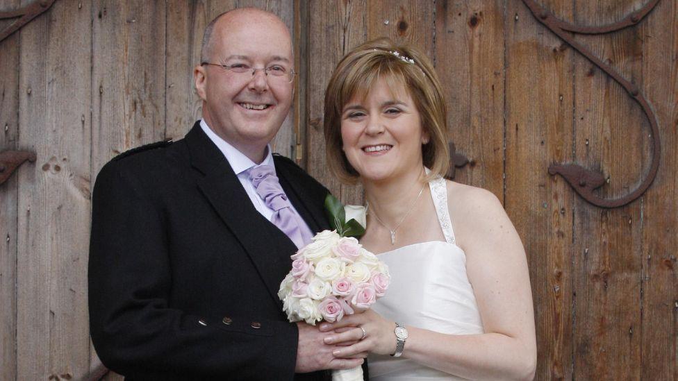 Nicola Sturgeon and Peter Murrell pictured on their wedding day in front of a wooden door. Sturgeon is wearing a white wedding dress, with Murrell wearing a dark jacket and purple tie. They are holding a bouquet of flowers.