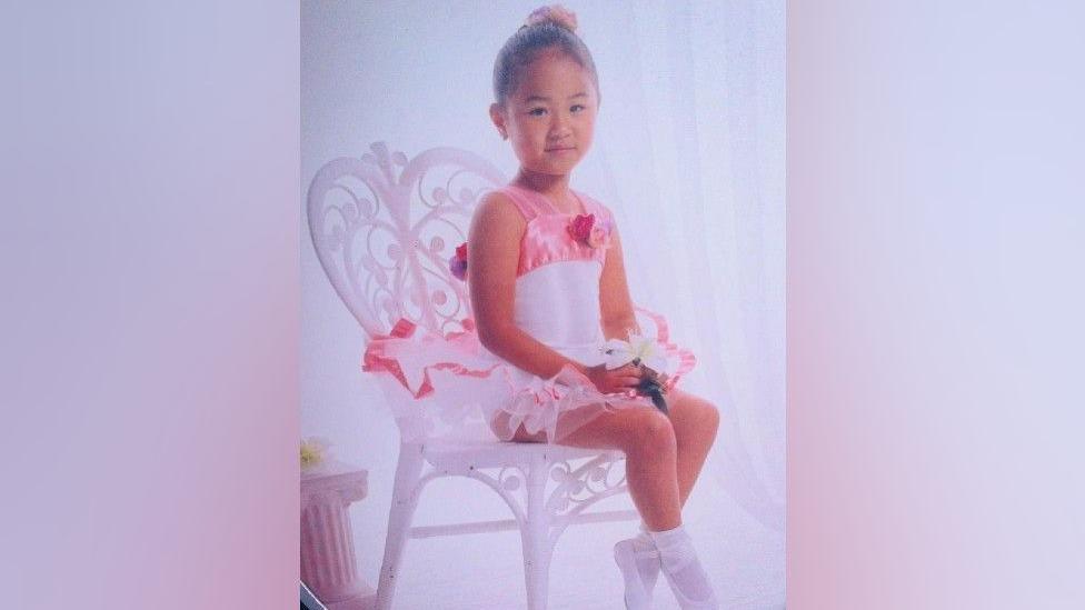 Jasmine as a child, sitting on a white chair. She is wearing a white leotard with baby pink straps and frills. She has her hair in a bun and a pink scrunchie. She has ballet shoes on her feet and is holding a white flower