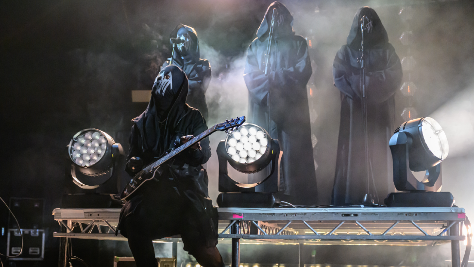 Four people on stage behind microphones, all dressed in black robes with hoods, and masks. There are also three lights on the stage.