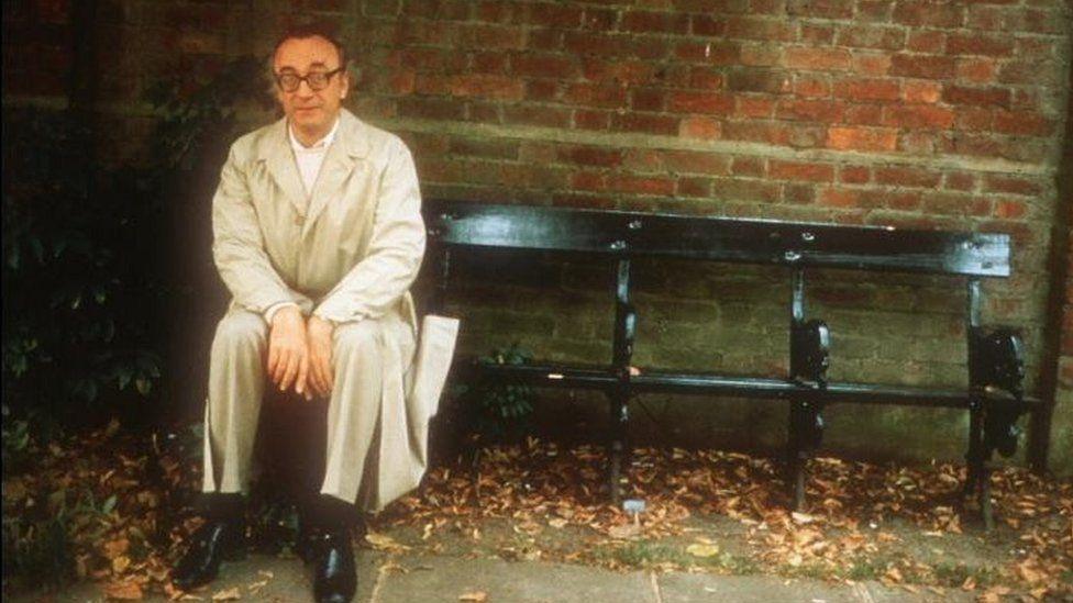 Alfred Brendel sitting on a park bench, wearing a trench coat