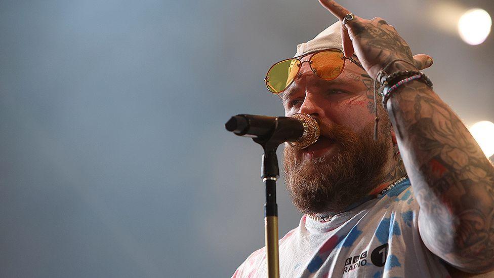 Teddy Swims wearing a cream cap and sunglasses resting on his forehead wearing a BBC Radio1 white t-shirt with blue and pink pattern performing at Radio 1's Big Weekend in July. 