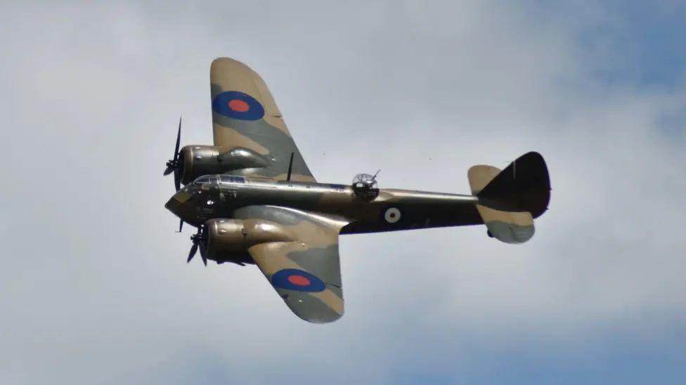 A world war 2 plan is pictured flying in the present day, presumably at an air show. The plane is in dark green camouflage colour, with a blue circle on each wing with a red circle inside.  