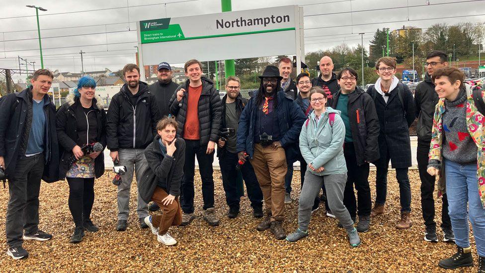 A group of men and women standing on gravel in front of a sign at Northampton railway station saying Northampton 