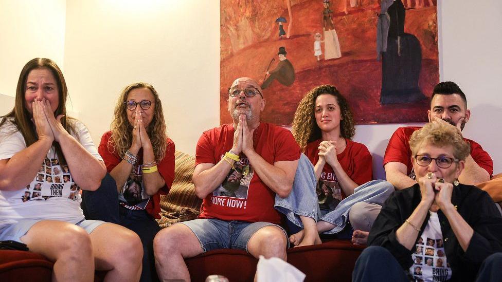 Relatives of Israeli hostage Eitan Horn, wearing red t-shirts with a picture of him that says "home now!", sit on a sofa and clasp their hands together as they watch a screen. They look hopeful and emotional