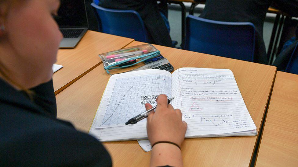 A view over the shoulder of a student who is writing in an exercise book containing handwritten text and drawings of graphs.