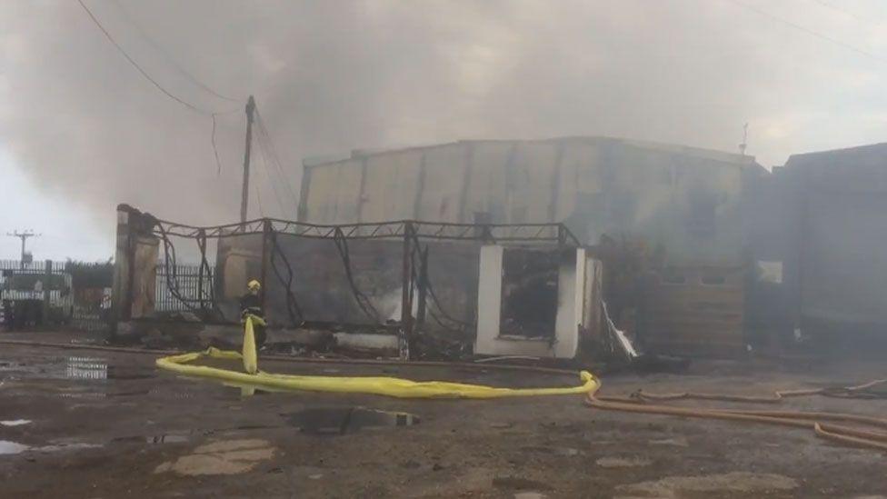 A firefighter by a burnt out industrial estate surrounded in thick smoke.