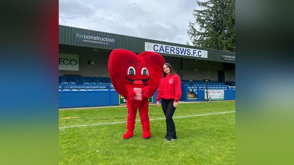 Faith has long brown hair and is standing on the grass of a football pitch wearing a red British Heart Foundation hoodie. She is standing next to a person that is dressed in a full-body red heart costume with a British Heart Foundation logo on it.