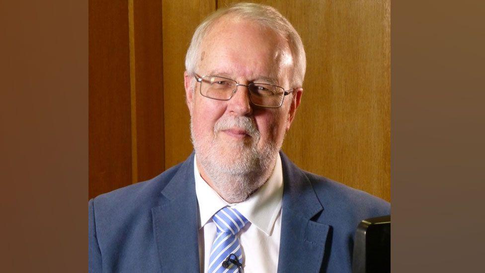 Ian Morton-Smith, with grey hair and a close-cropped grey beard, wearing metal-framed glasses, a blue jacket, white shirt and blue-striped tie