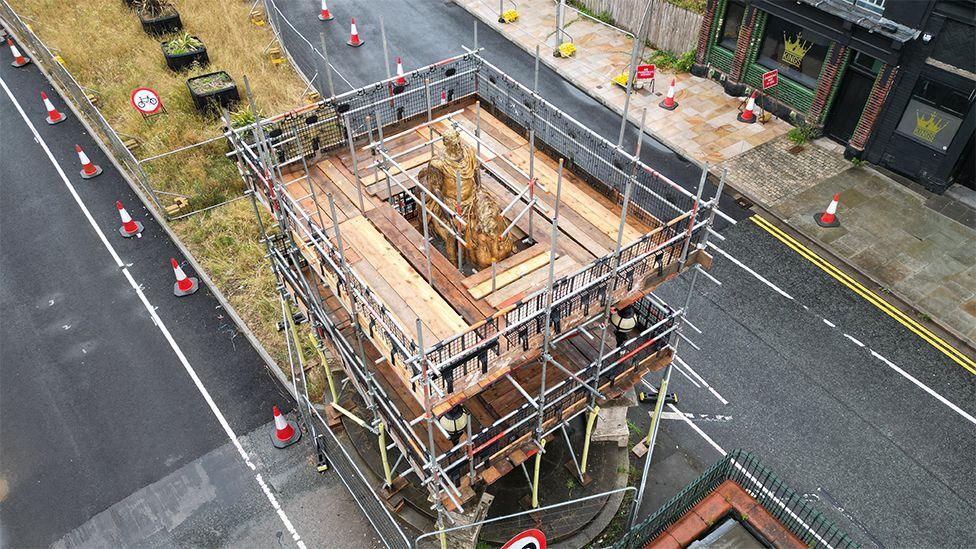 An aerial view of the scaffolding around the statue of King William III on his golden horse. It is surrounded by a platform of wooden planks. There are traffic cones along the road on both sides.