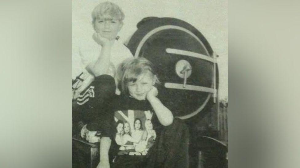 A black and white image of a news paper of a young boy and a young girl posing in front of a miniature train.