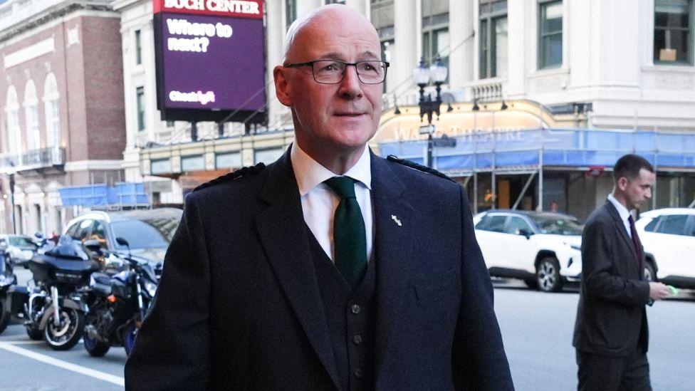 John Swinney, who is bald and wearing glasses, walks on a city street in formal wear 