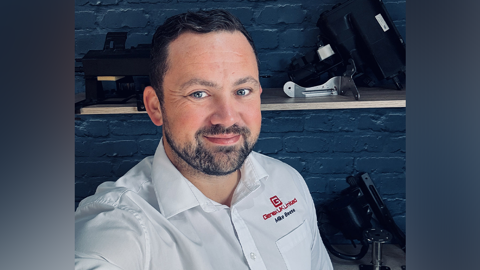 Michael Beese pictured taking a selfie against a dark grey brick wall. He has short black hair and a dark stubble beard. He is wearing a white collar shirt with the red Genex UK Limited logo.