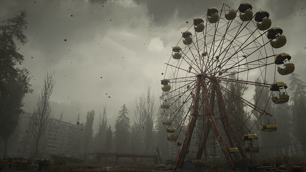 A screenshot from Stalker 2 shows a ferris wheel with obvious signs of decay and disrepair against a desolate backdrop of overgrown trees, a dilapidated block of flats and various ground debris. A foreboding grey mist covers the scene and specks of black ash fall from the sky.