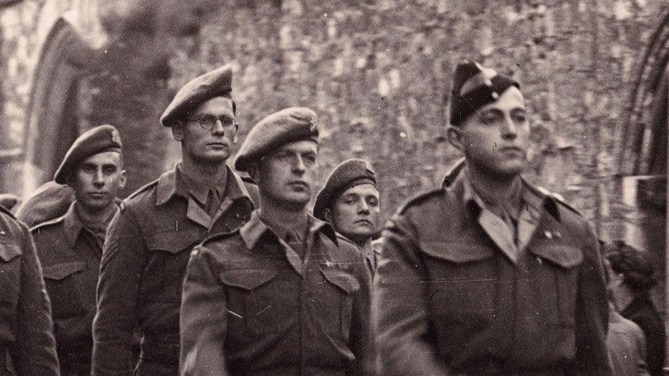 A black and white image showing men from the Cambridgeshire Regiment marching past Ely's war memorial in late 1945. They are in Army uniform. They are being watched by men, women and children.