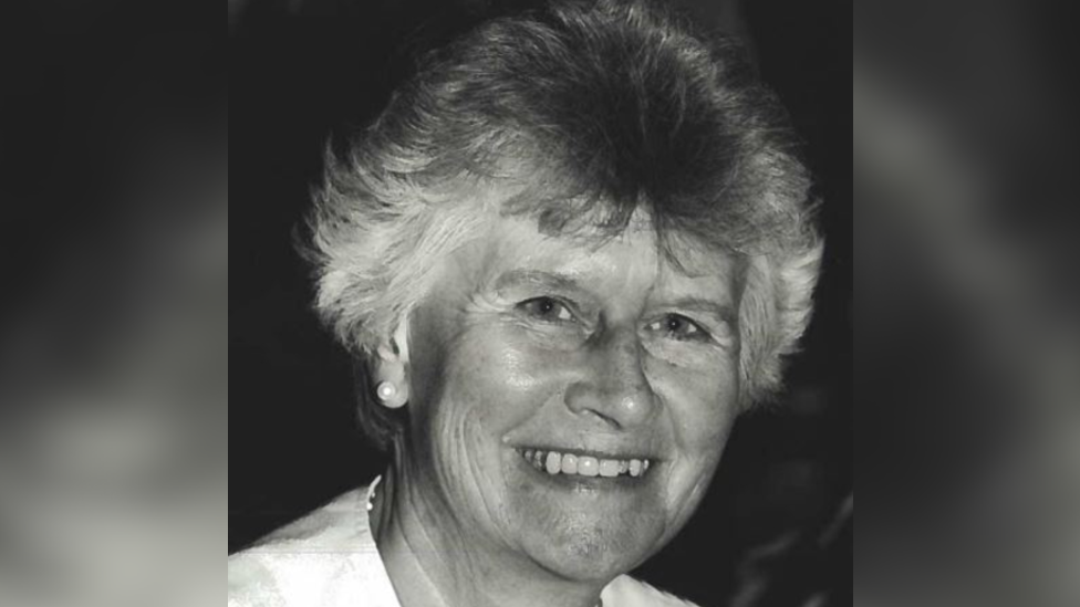 A black and white portrait photo of Beryl Purdy. She has short grey hair and is smiling at the camera. She is wearing a white top and pearl earrings.