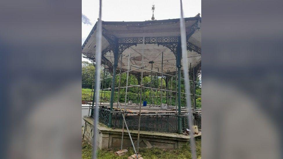 The bandstand photographed up-close from behind a fence. The structure is covered in scaffolding. There are trees in the background.