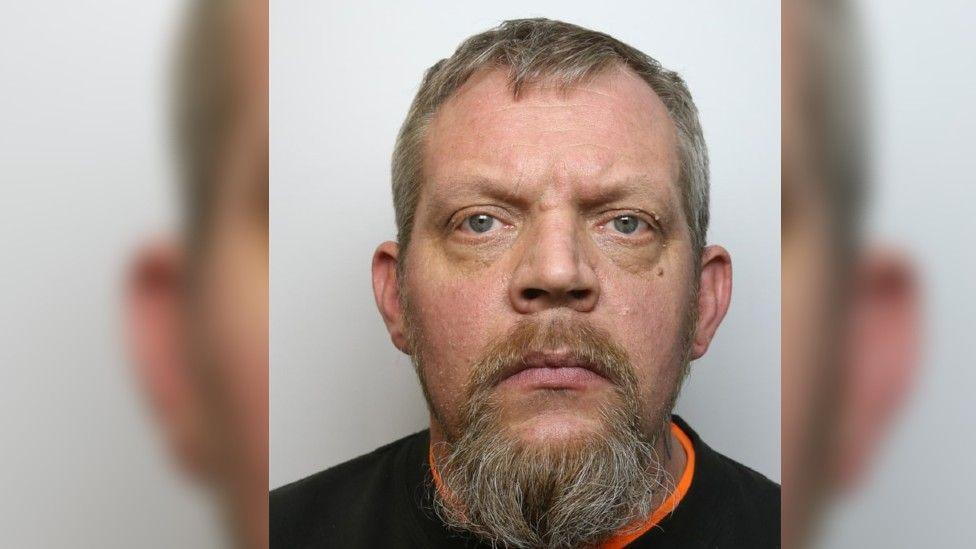 Mugshot of Stephen Burrows who has a beard and is wearing a black top with an orange rim.