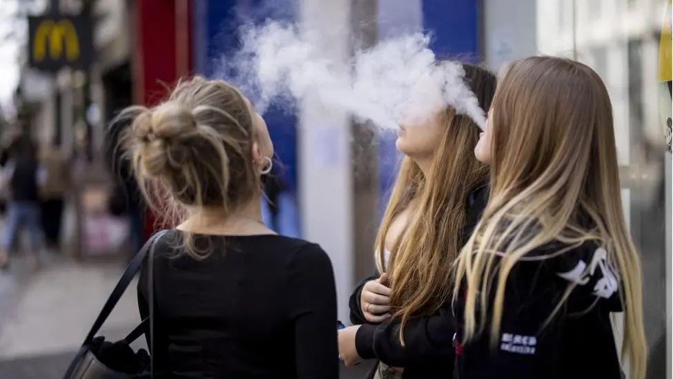 Three young women with long hair are standing in a street using vapes. They are exhaling plumes of vapour which looks like white smoke.