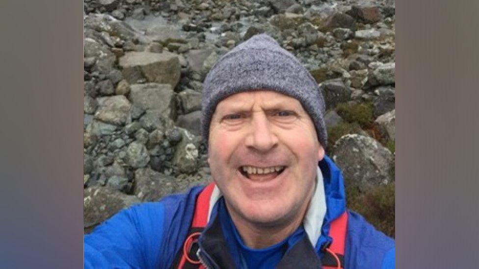 Neil McGibbon taking a selfie, wearing a grey hat on a rocky landscape