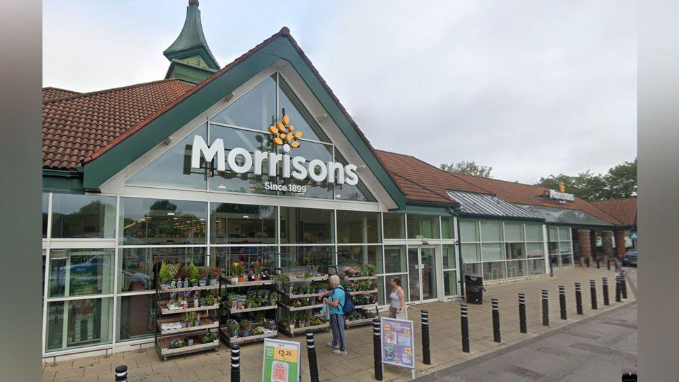 The front entrance of Morrisons, with a large sign on the triangle shaped front.
It has a red tiled roof and green edging, and glass front. Stands of plants are outside the front, which has a paved area lined with bollards. Two people stand looking at the plants.