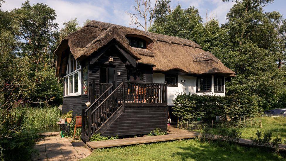 The waterside chalet has stairs leading up to it. It has black wooden cladding on one side and pained white on the other. It has a thatched roof. 