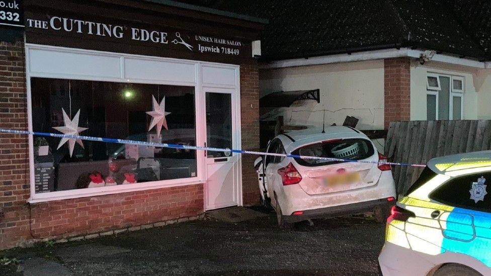 A white Ford Focus can be seen stuck between a bungalow and hair salon. You can see the back of the car which is wedged between the buildings. A police car is also at the scene and a blue and white police tape cordon is also in place. 