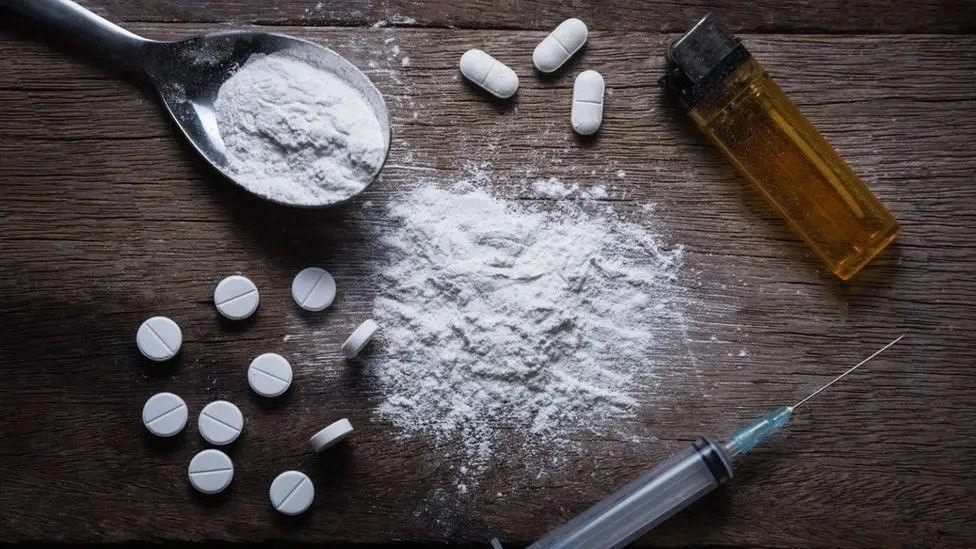 Pills laid out on a wooden table, alongside a crushed white powder, a spoon of white powder, a syringe and yellow lighter