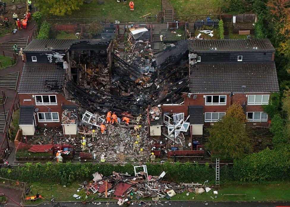 A drone shot of row of houses with the central house collapsed in a pile of rubble and burnt wood. Emergency service workers in bright orange overalls are at the centre of the rubble. October 16, 2024. 