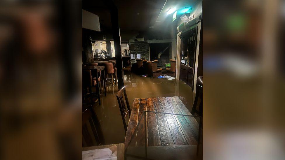 Dark photo of a flooded pub. Dark murky water is about a few inches high and you can see the bar at the back.