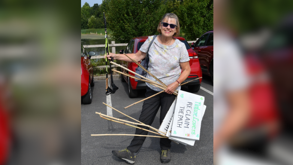 Pat stands holding a walking pole and signs that read 'Reclaim the Path'. She is standing in a carpark and is wearing a white top with walking trousers and walking boots.