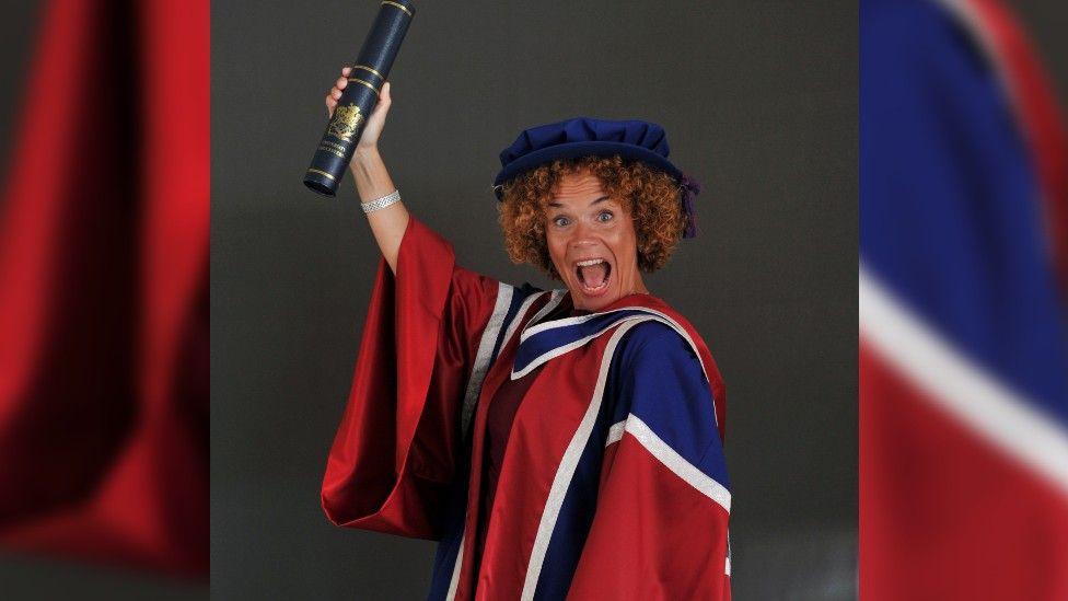 Sabrina Pace-Humphreys in graduation robes of red, white and blue, with a navy graduation cap covering her curly hair, and holding her graduation certificate in a case, up in the air with her right hand.