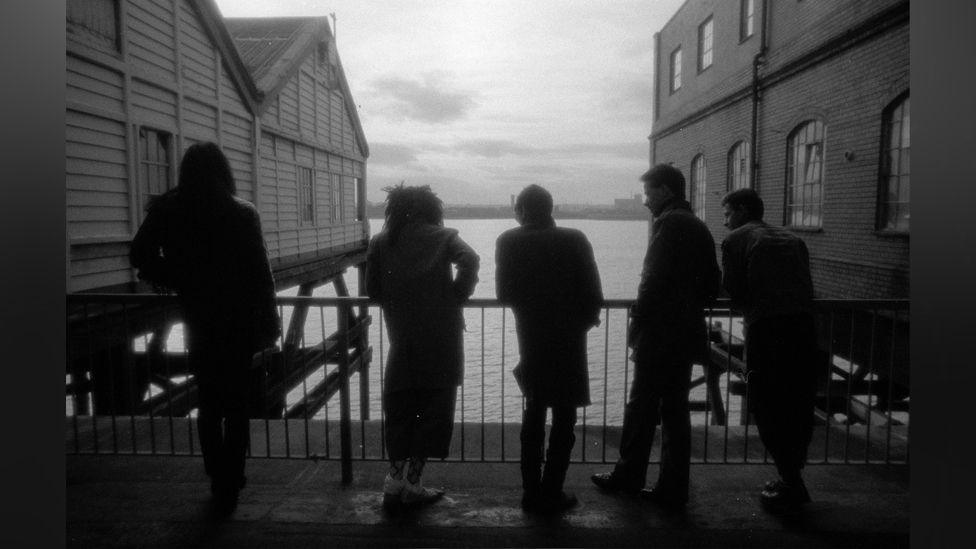 Black and white photograph of five silhouetted men leaning on railing looking out towards river with dock buildings on either side