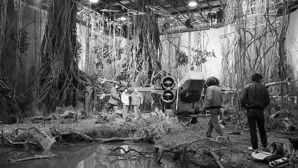 A black and white image of a Star Wars set, which looks like a swamp with large trees and creepers draped around a pond. Some people stand on the set and there is a plane or spaceship in the middle.