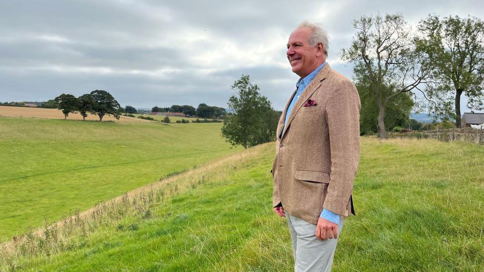 A man in a tweed jacket an blue shirt looks out over a sloping grass field with trees in the background