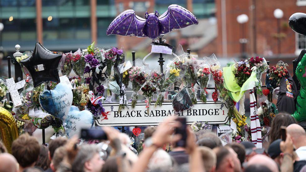 A Black Sabbath Bridge sign with numerous tributes stuck to the railings, a bat ballon above them, and the heads of the crowd in the foreground
