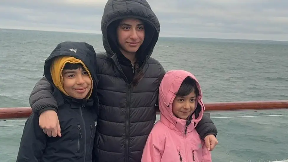 Three siblings stand together posing for a photo - Hussam on the left, Rahaf in the middle with her arms around her brother and Sara, on the right.  They are smiling and all have the hoods of their coats up. They are standing against the backdrop of the sea.