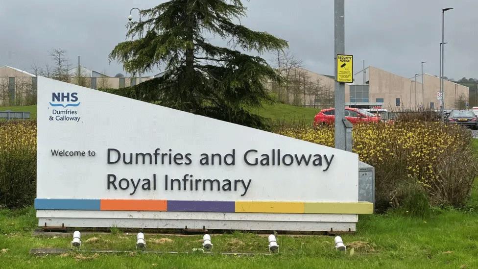 A sign reading welcome to Dumfries and Galloway Royal Infirmary in front of a tree and some lamp posts leading up to some hospital buildings in the distance