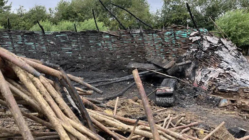 The charred remains of a wicker round structure and scorched earth, with trees in the background.