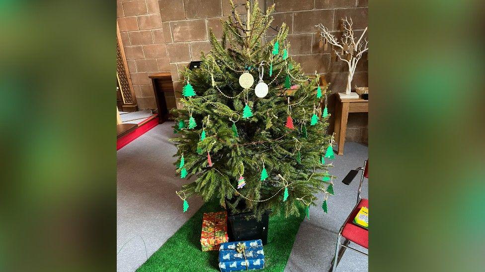 Medium-sized Christmas tree with white beads draped on it, Christmas tree-shaped baubles and some presents at the bottom.