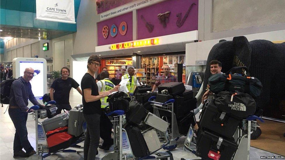 A group of five men standing in a busy airport terminal. There is a sign that says 'Cape Town' above them. There is a shop in the background and a large model elephant to the right.
